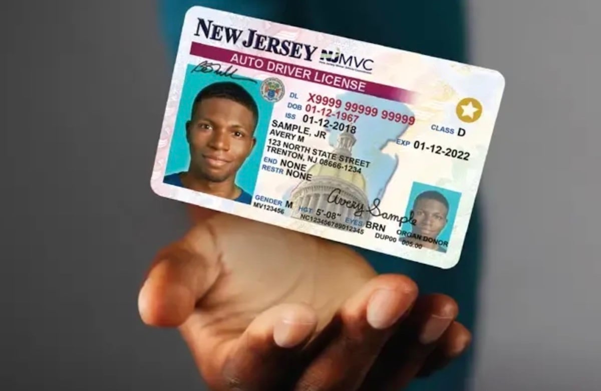 A hand holds a New Jersey auto driver license featuring a photo of a man, with sample personal information and a visible gold star in the top right corner of the card.