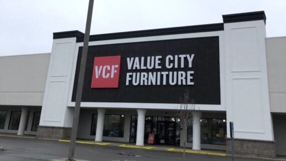 The image shows the storefront of a Value City Furniture store with a large black sign featuring the red VCF logo and white lettering above the entrance. The building has white pillars and a mostly empty parking lot.