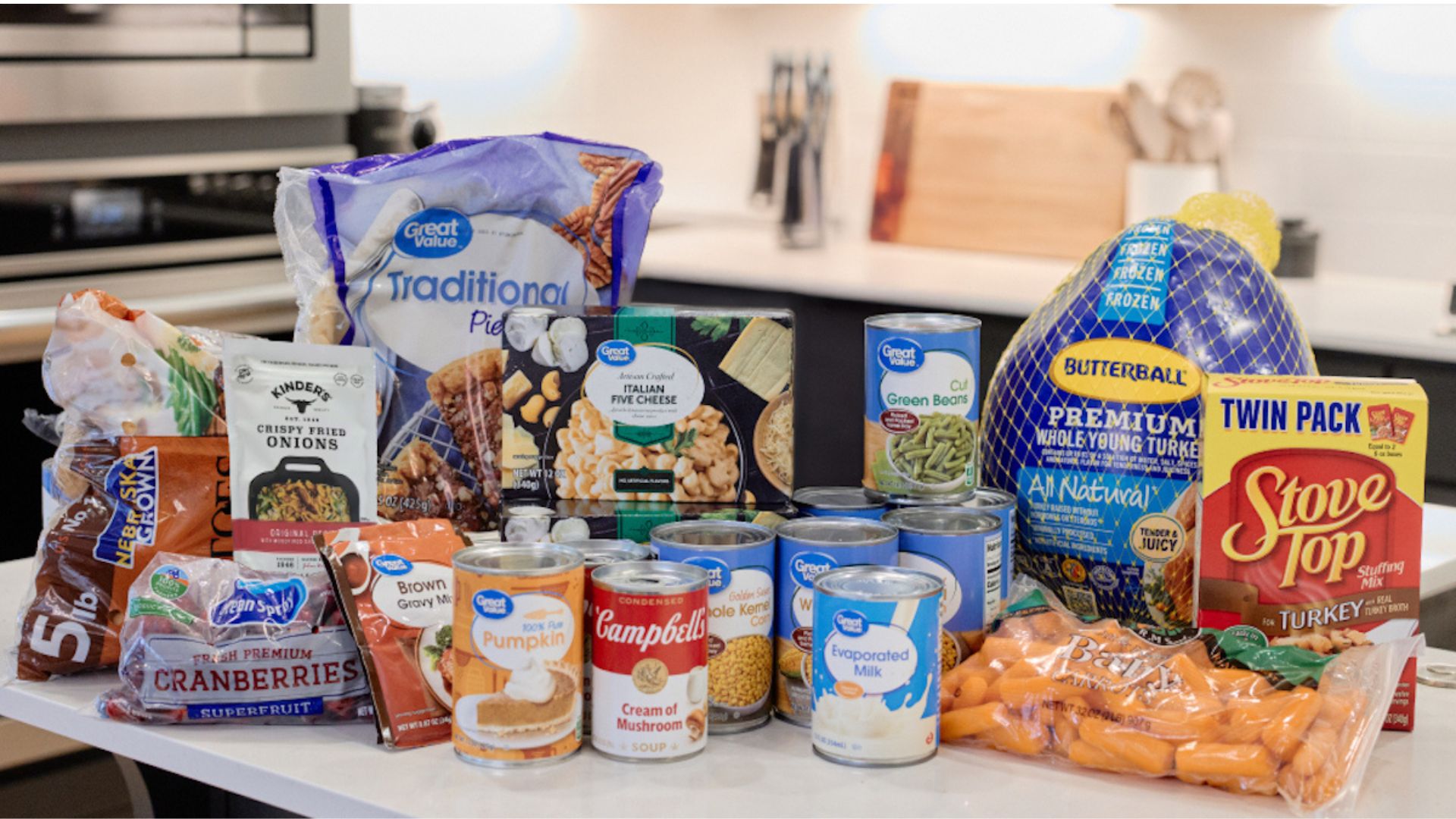 A variety of Thanksgiving food items on a kitchen counter, including a frozen turkey, canned vegetables, pie crusts, boxed stuffing, canned pumpkin, cheese, onions, carrots, and bags of cranberries and potatoes.