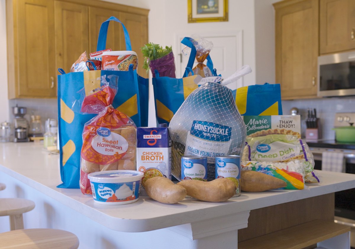 A kitchen island with grocery bags and various Thanksgiving food items, including a turkey, canned goods, sweet potatoes, sweet Hawaiian rolls, chicken broth, whipped topping, and a frozen pumpkin pie.
