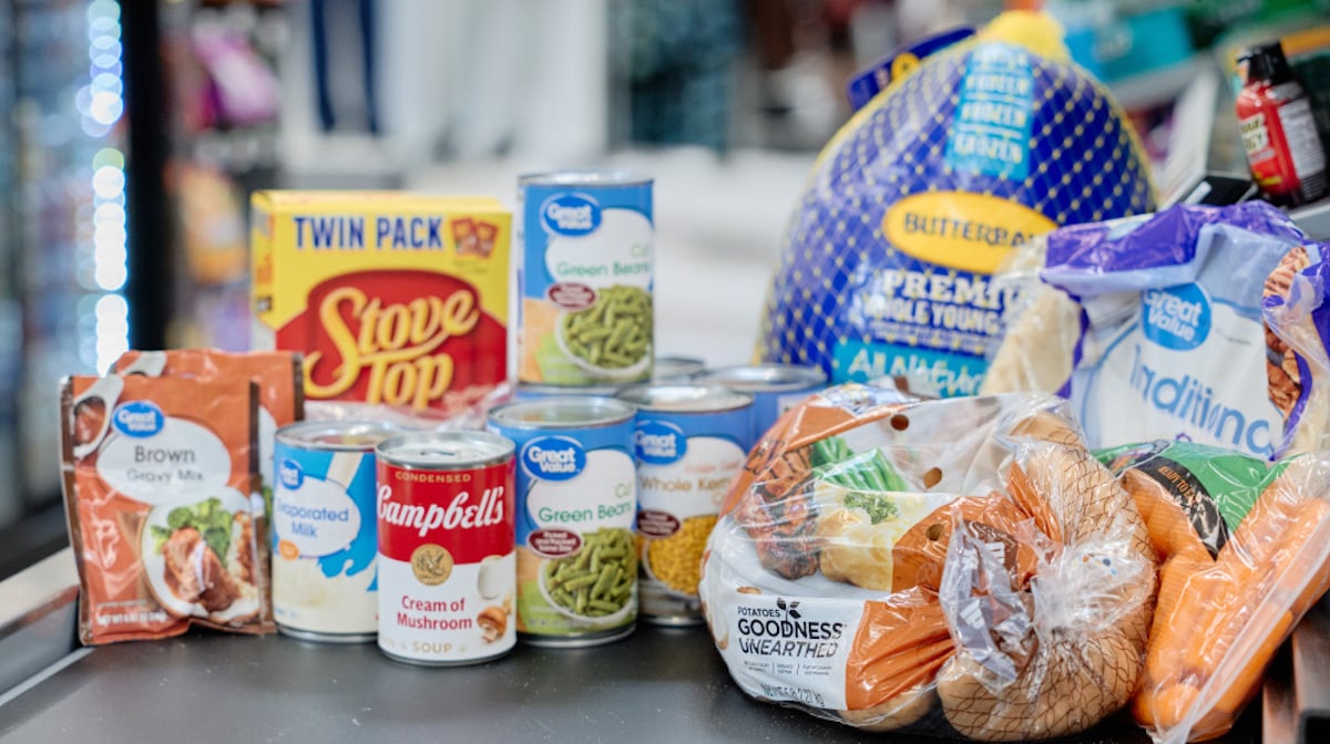 A grocery store checkout conveyor belt with canned green beans, stuffing mix, gravy mix, condensed milk, mushroom soup, a whole turkey, potatoes, onions, and a loaf of white bread.