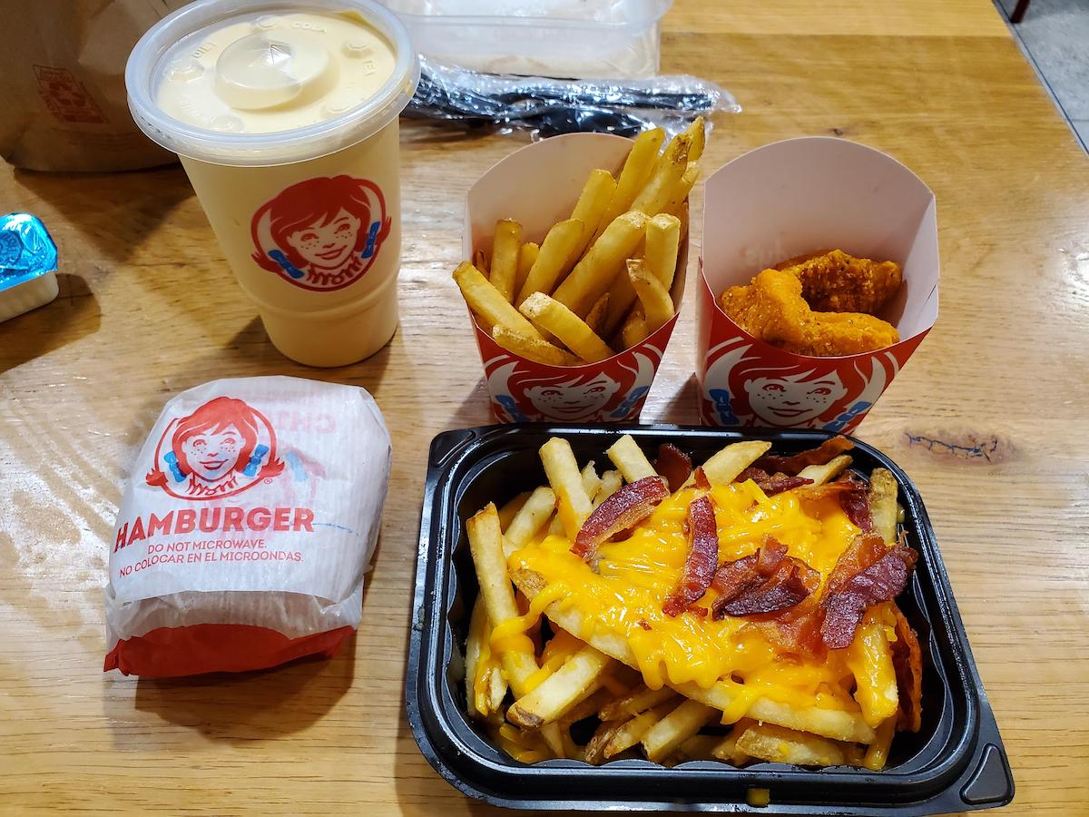 A fast food meal with a wrapped hamburger, two servings of fries (one with melted cheese and bacon), chicken nuggets, and a cold drink on a wooden table, all in Wendy’s branded packaging.
