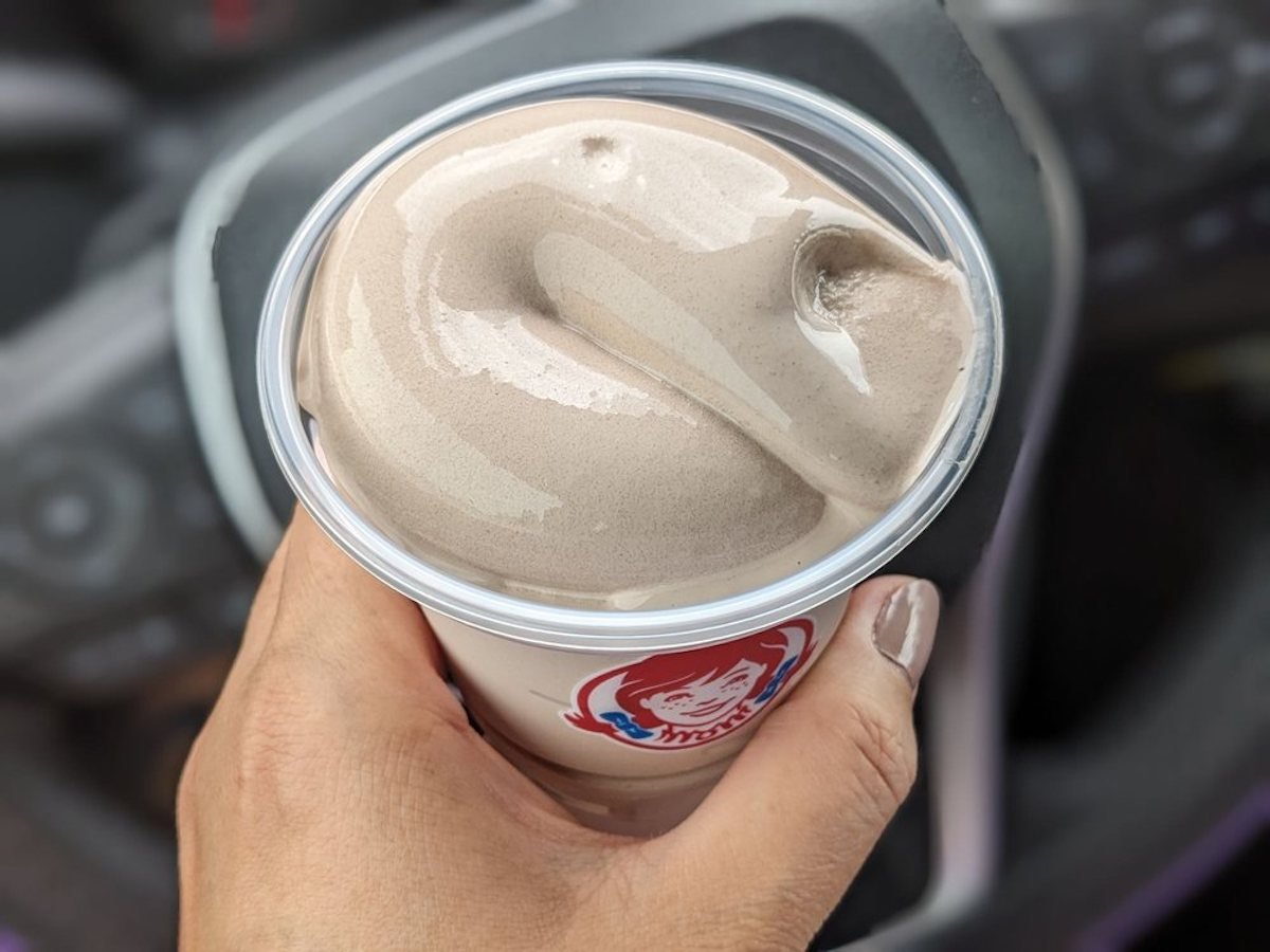 A hand holding a cup of Wendy's chocolate Frosty with a smooth, swirled top, against a blurred car interior background.
