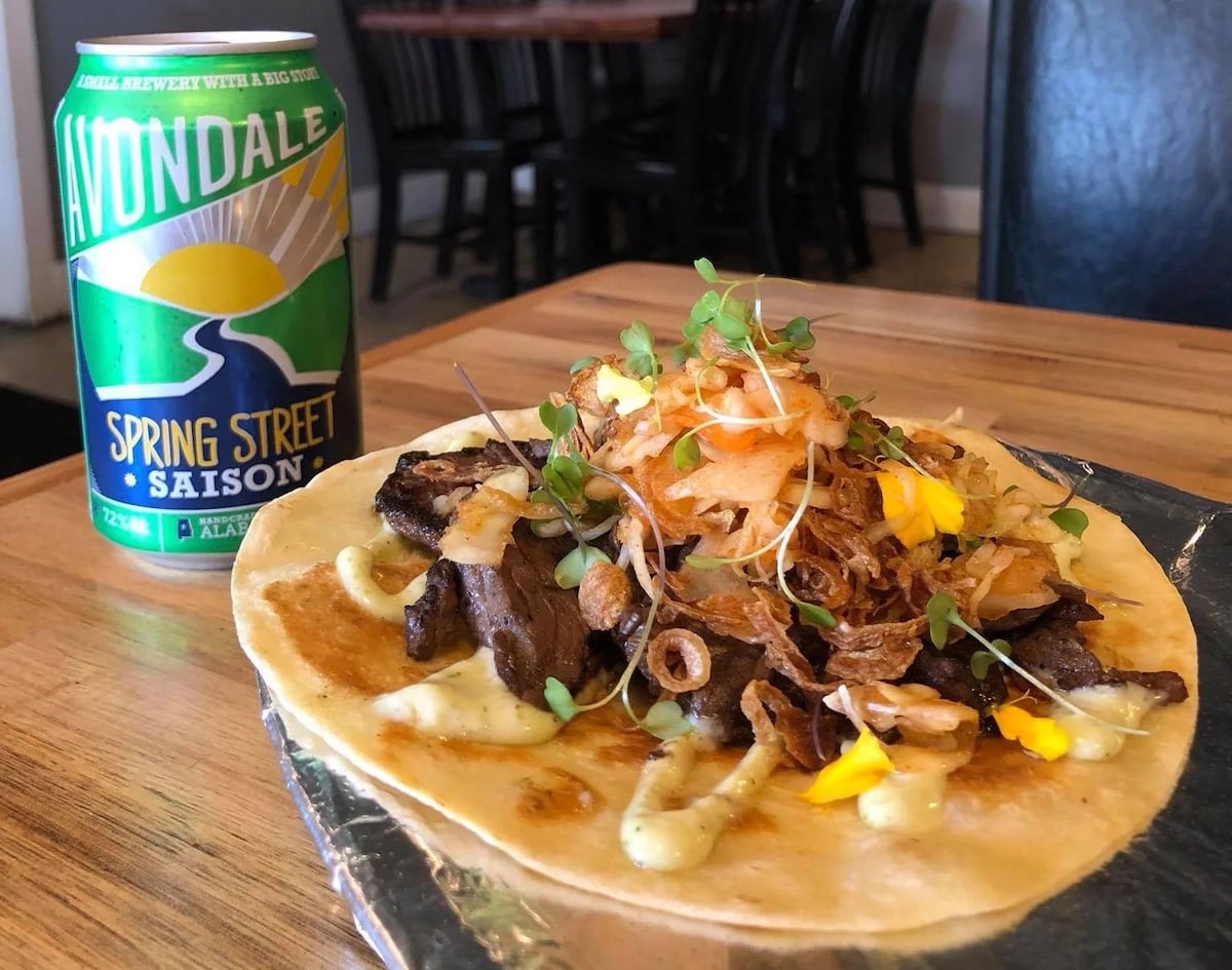 A taco filled with grilled meat, microgreens, fried onions, and sauce sits on foil on a wooden table beside a can of Avondale Spring Street Saison beer—an inviting find at one of the city's best under-the-radar restaurants.
