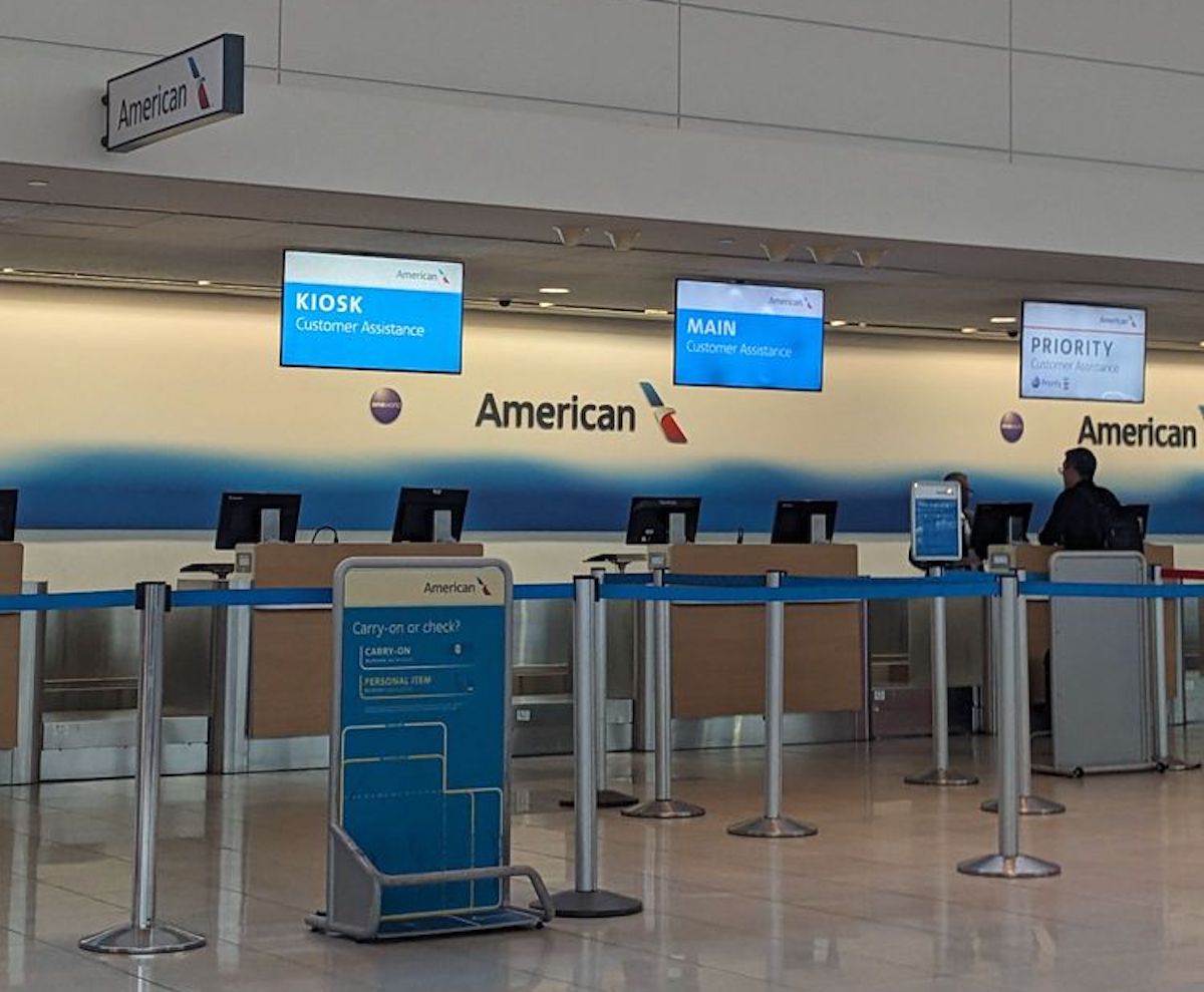 American Airlines check-in counters at an airport, with digital signs for kiosk, main, and priority customer assistance. A sign with baggage information stands in the foreground; the counters are mostly empty.
