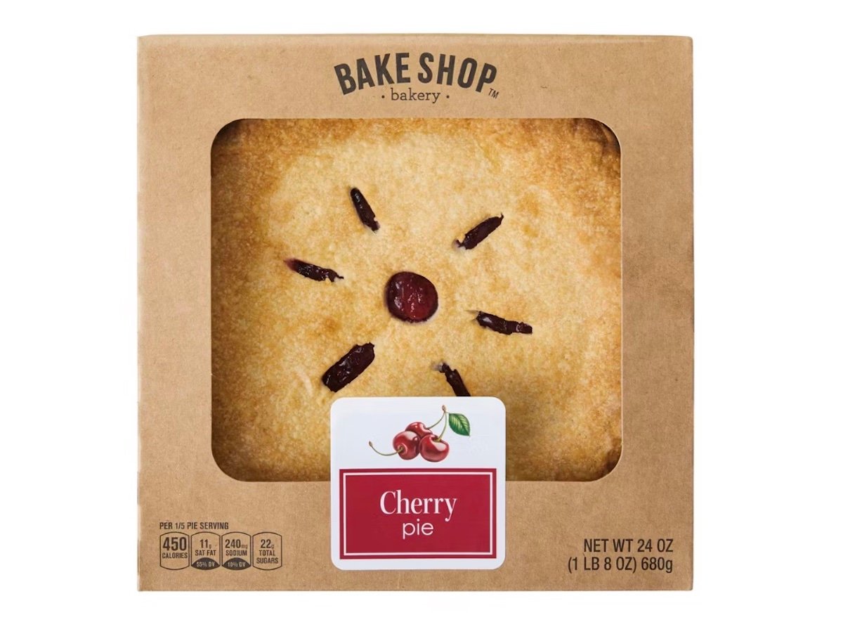 A boxed cherry pie from Bake Shop Bakery, shown through a clear window in the packaging. The golden-brown crust has cherry filling peeking through slits, and a label on the box reads "Cherry pie" with an image of cherries.