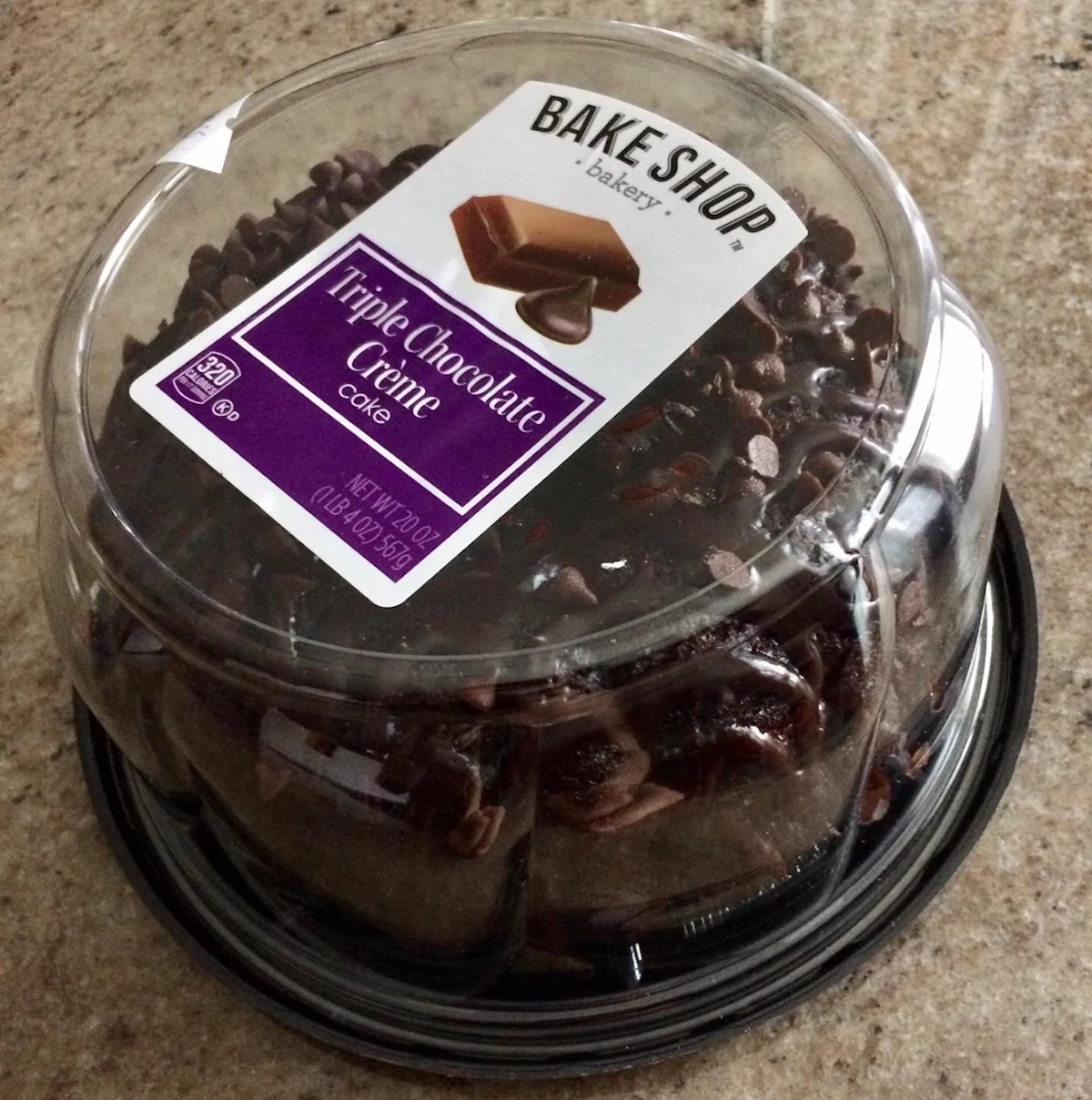A round triple chocolate crème cake with chocolate chips, packaged in a clear plastic container labeled "Bake Shop Bakery" on a tan countertop.