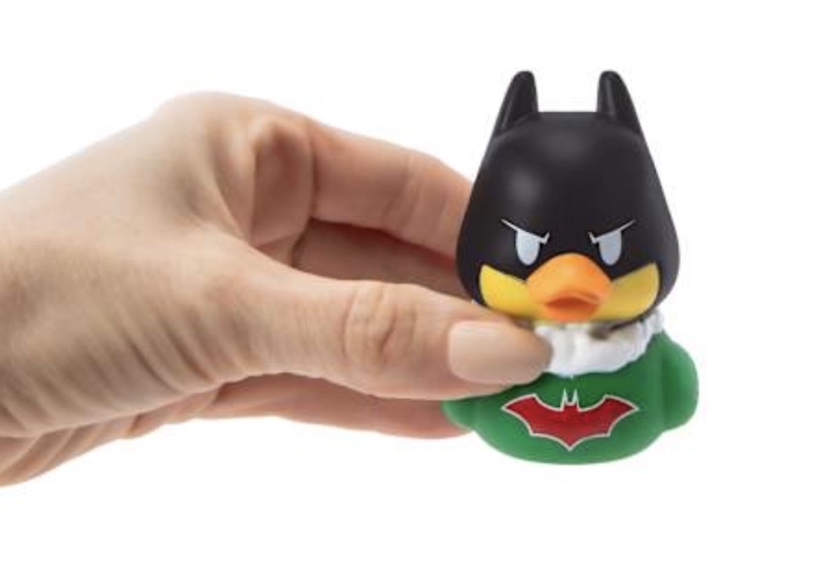 A hand holds a small rubber duck designed to look like a superhero, with a black mask, yellow beak, green suit, and red bat symbol on its chest.