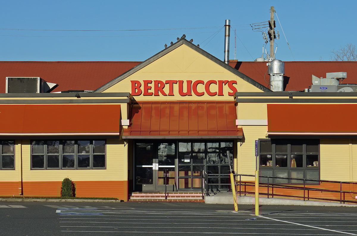 A Bertucci’s restaurant exterior with large red letters on the building, glass doors, and windows. There is a ramp and empty parking spaces in front, under a clear blue sky.