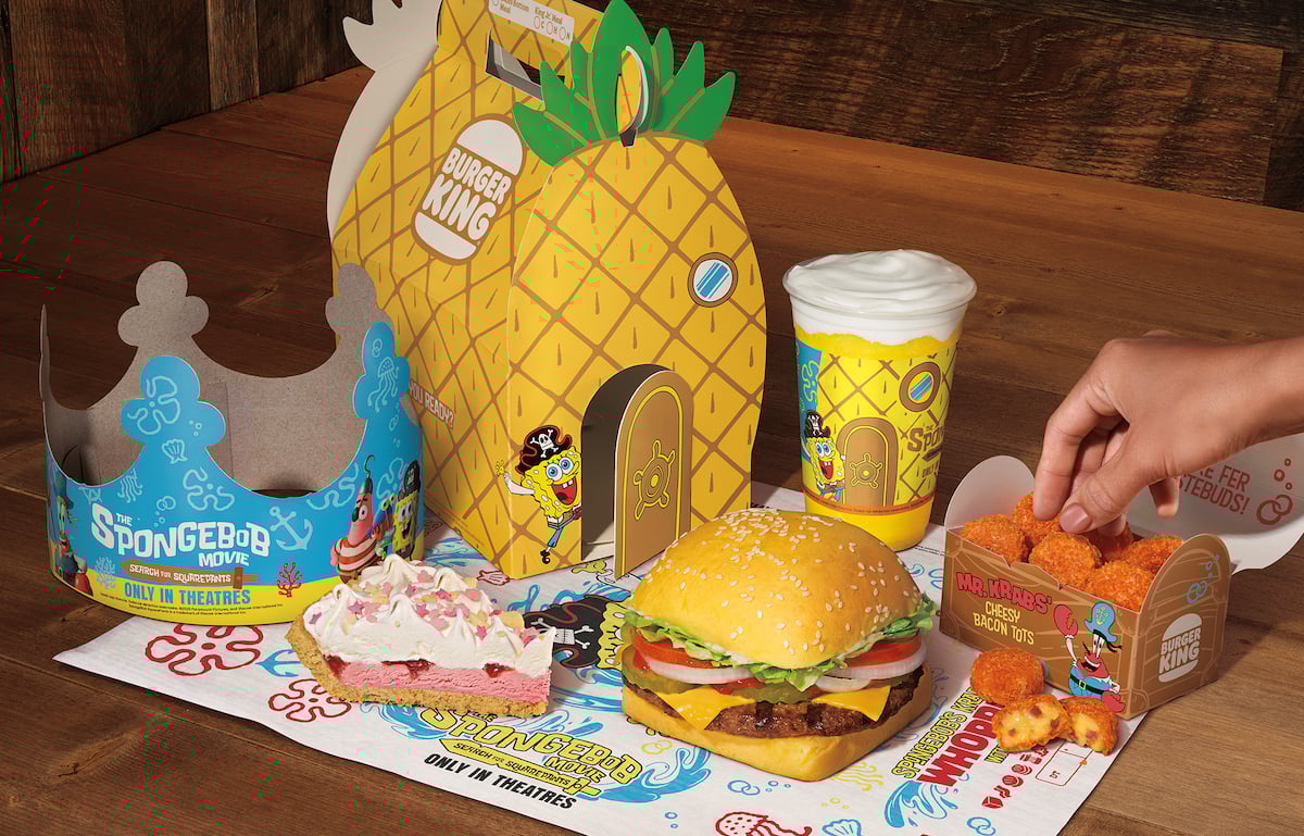 A Burger King meal with a Spongebob Squarepants theme, including a burger, fries, drink, dessert, a cardboard crown, and a pineapple-shaped box, with a hand picking up a piece of food.