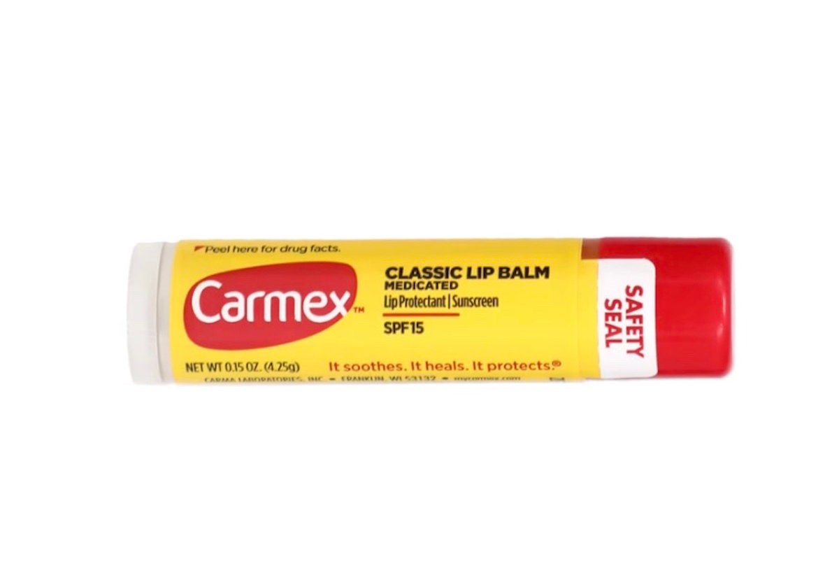 A Carmex Classic Lip Balm tube with a white cap and a yellow-and-red label featuring SPF 15, medicated, and safety seal details. The text reads, "It soothes. It heals. It protects.