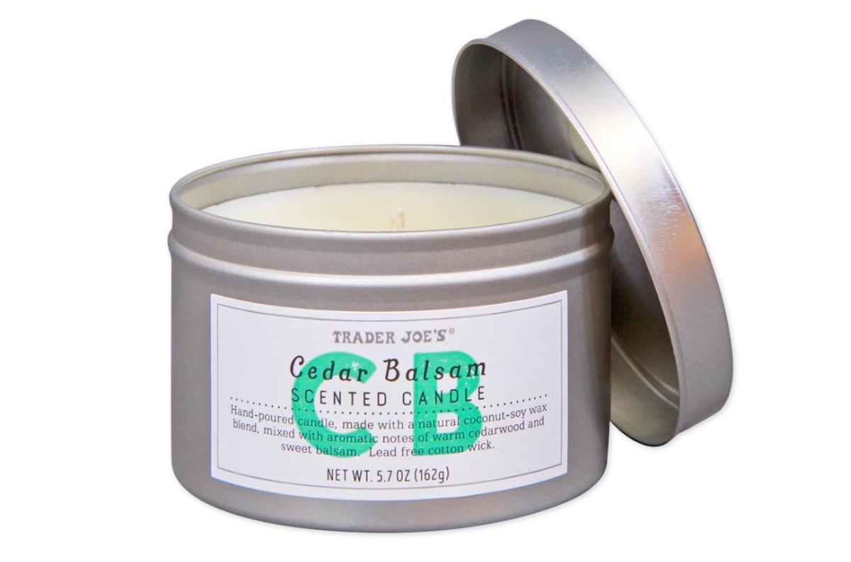 A silver tin of Trader Joe’s Cedar Balsam Scented Candle with the lid partially off, revealing a white candle inside. The label describes a coconut-soy wax blend with cedarwood and sweet balsam scents.