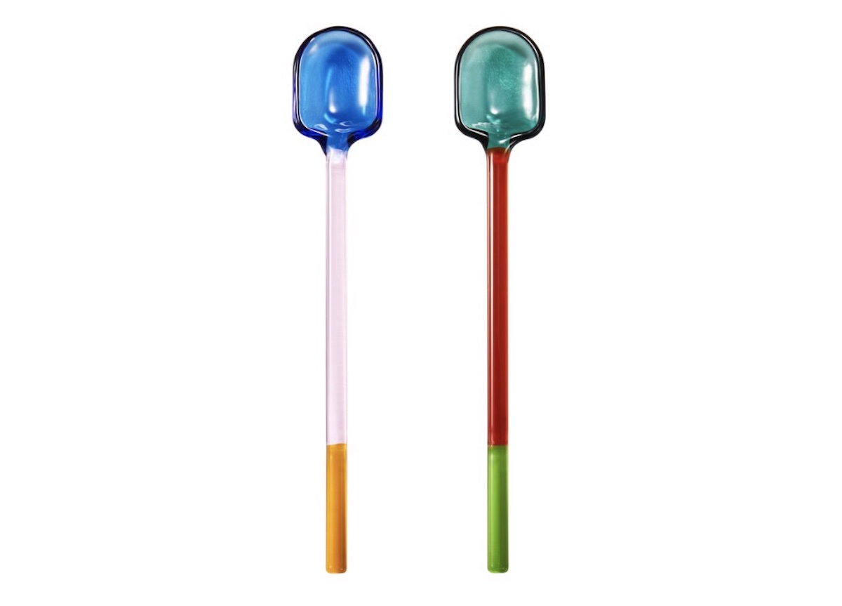 Two colorful glass spoons with rounded heads, one with a blue top and pink-orange handle, the other with a green top and red-green handle, both against a white background.