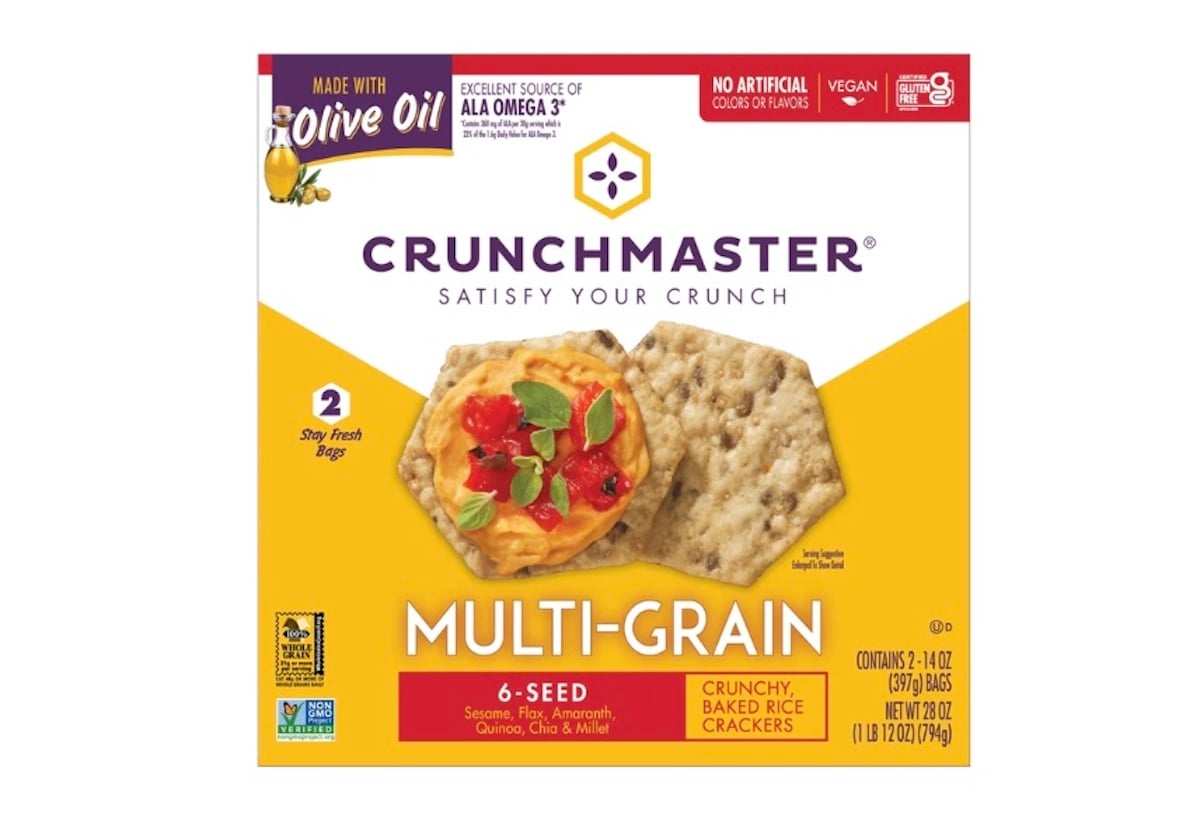 Box of Crunchmaster Multi-Grain 6-Seed crunchy baked rice crackers, featuring an image of crackers topped with spread and vegetables. The box highlights being made with olive oil and is labeled vegan.