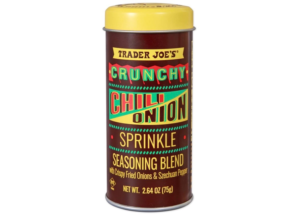 A cylindrical container of Trader Joe’s Crunchy Chili Onion Sprinkle Seasoning Blend with a yellow lid and colorful text on a brown label. The net weight is 2.64 ounces (75g).