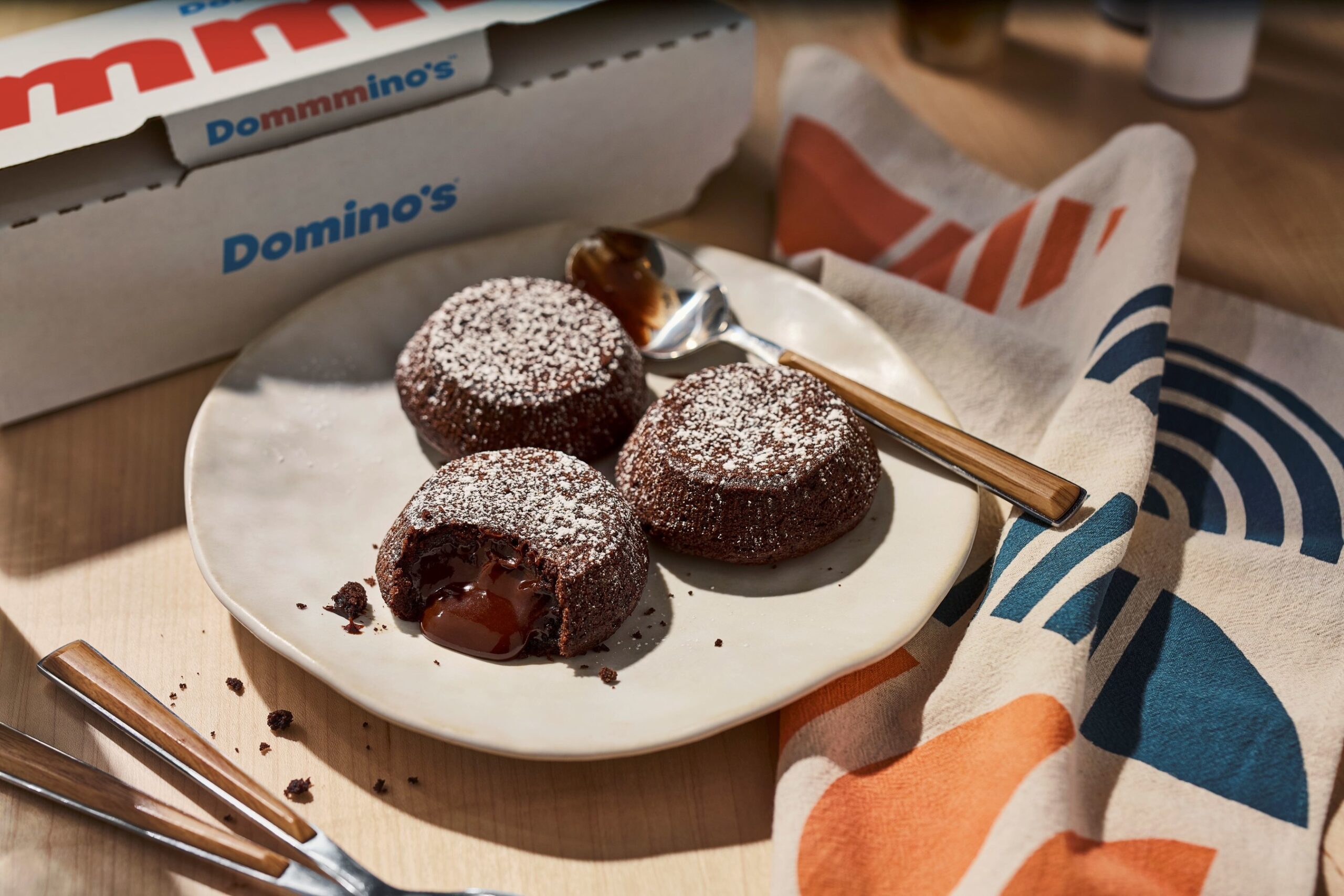 Three chocolate lava cakes dusted with powdered sugar are served on a plate, with one cut open to reveal melted chocolate inside. A spoon rests beside the plate, and a Domino’s pizza box is seen in the background.
