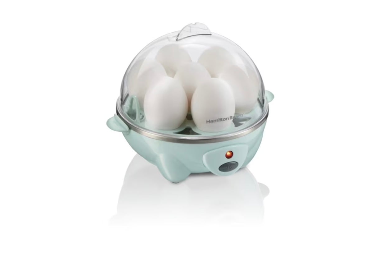 A light blue electric egg cooker with a transparent dome lid, holding seven white eggs. A small indicator light and button are visible on the front of the device.