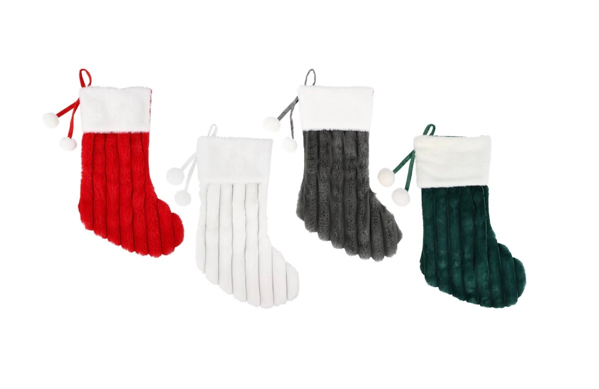 Four plush Christmas stockings in red, white, gray, and green, each with white cuffs and two pom-poms hanging from a ribbon, are arranged in a row against a white background.