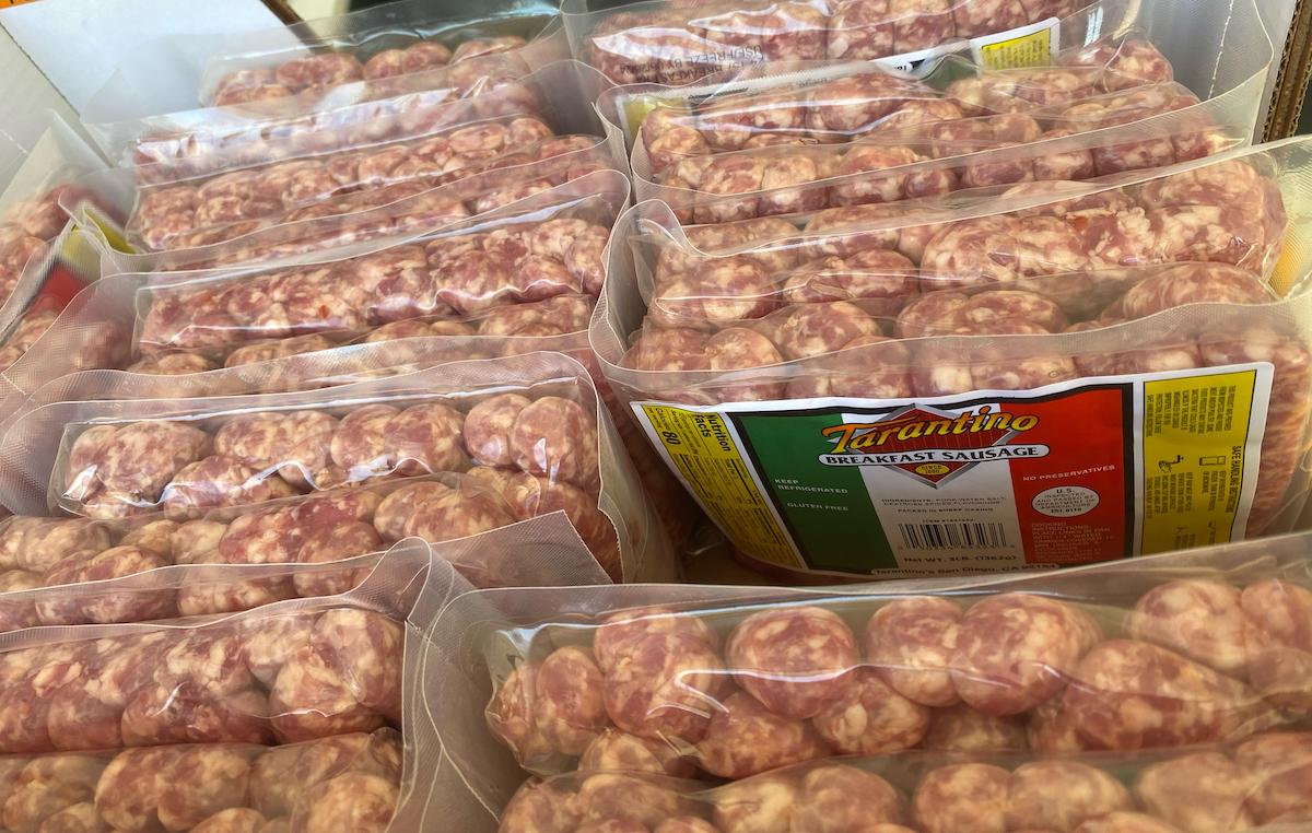 Packages of Tarantino breakfast sausages are stacked in a box. The sausages are sealed in clear plastic, and one package displays the brand name and a label with red, green, and white colors.