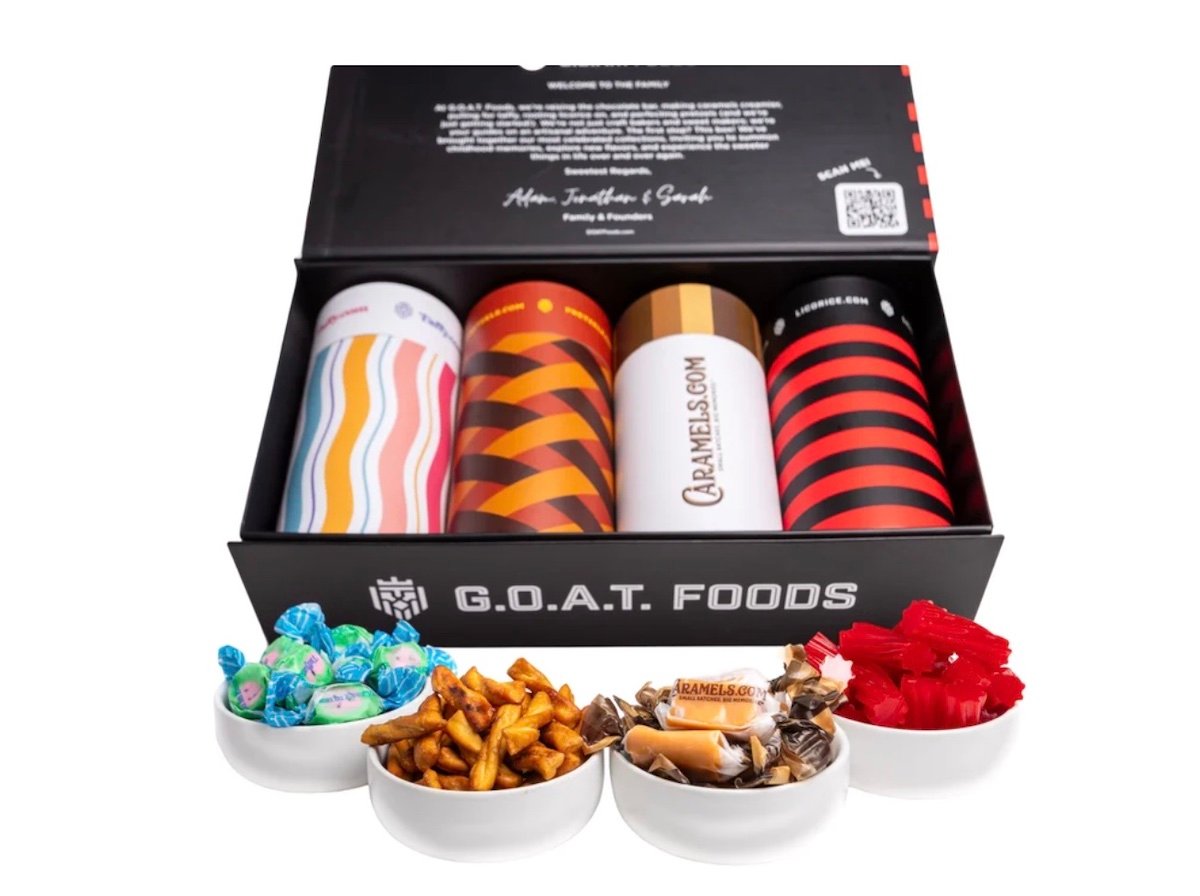 An open G.O.A.T. Foods box displays four colorful canisters and five small bowls filled with assorted candies, including green-wrapped sweets, pretzel sticks, caramels, and red licorice.