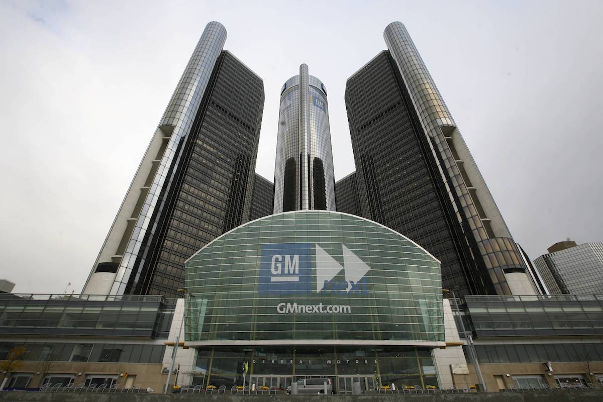 A wide-angle view of the GM Renaissance Center in Detroit, featuring four tall glass towers surrounding a central cylindrical skyscraper with GM logos and a large glass entrance in front.