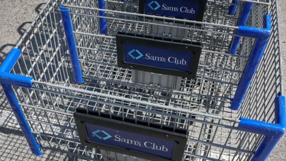 Several metal shopping carts with blue handles, stacked together outside. Each cart has a sign that reads "Sam's Club" with the store's logo displayed on the front panel.