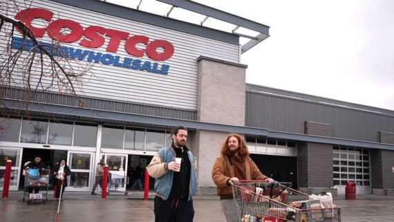 Two people with a shopping cart walk outside a Costco Wholesale store. One holds a drink while the other pushes the cart filled with groceries. The Costco sign is visible above the entrance. Other shoppers are in the background.
