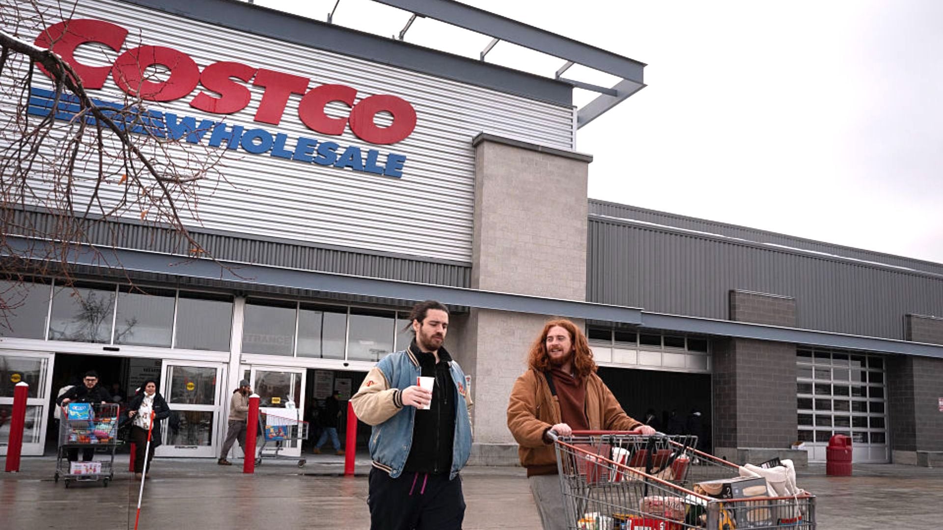 Two people with a shopping cart walk outside a Costco Wholesale store. One holds a drink while the other pushes the cart filled with groceries. The Costco sign is visible above the entrance. Other shoppers are in the background.