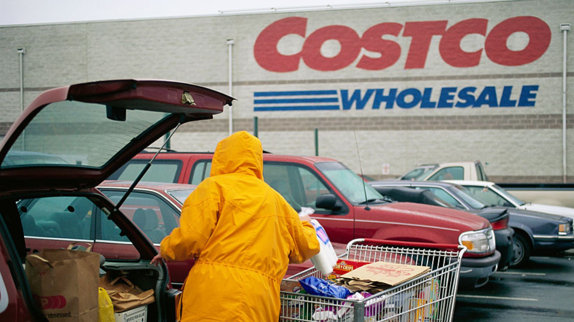 A person in a yellow raincoat loads groceries from a full shopping cart into the trunk of a red car in a parking lot outside a Costco Wholesale store. Other cars are parked nearby.