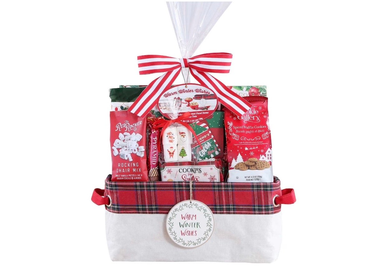 A festive holiday gift basket wrapped in clear plastic with a red-and-white striped bow, filled with cookies, cocoa mix, and treats, in a plaid-trimmed container labeled "Happy Winter Wishes.