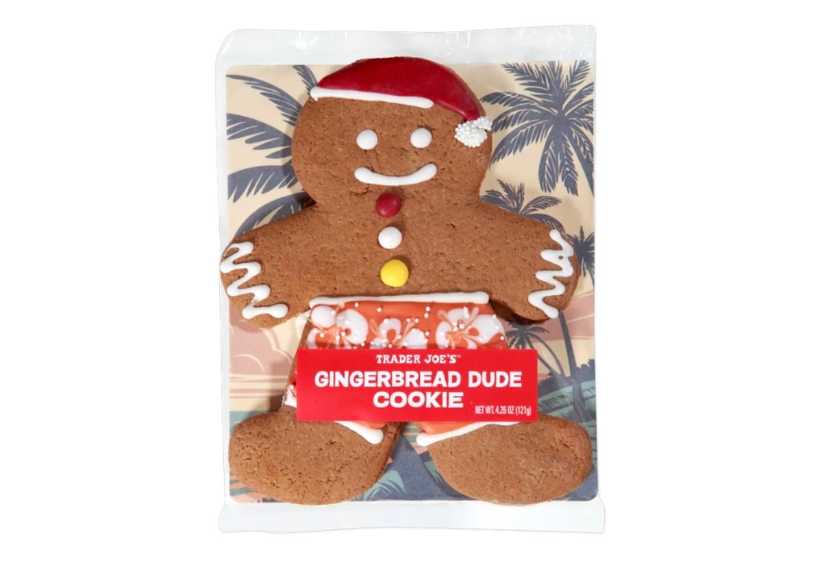 A Trader Joe’s gingerbread cookie shaped like a person, decorated with a Santa hat, red and yellow buttons, and white icing. The cookie wears orange floral swim trunks and is packaged in front of a tropical palm tree background.