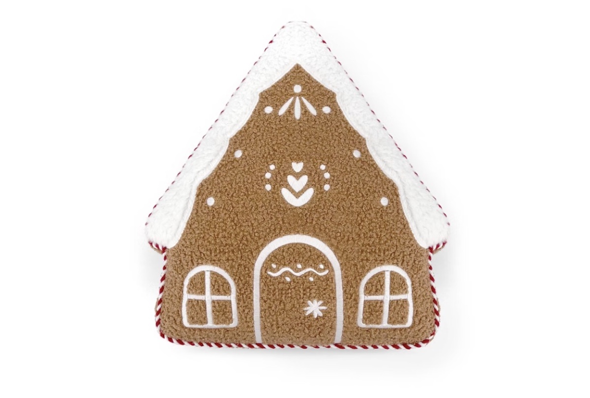 A plush toy shaped like a gingerbread house with white icing details, including a door, windows, and decorative patterns, and topped with white “snow” along the roof edges.