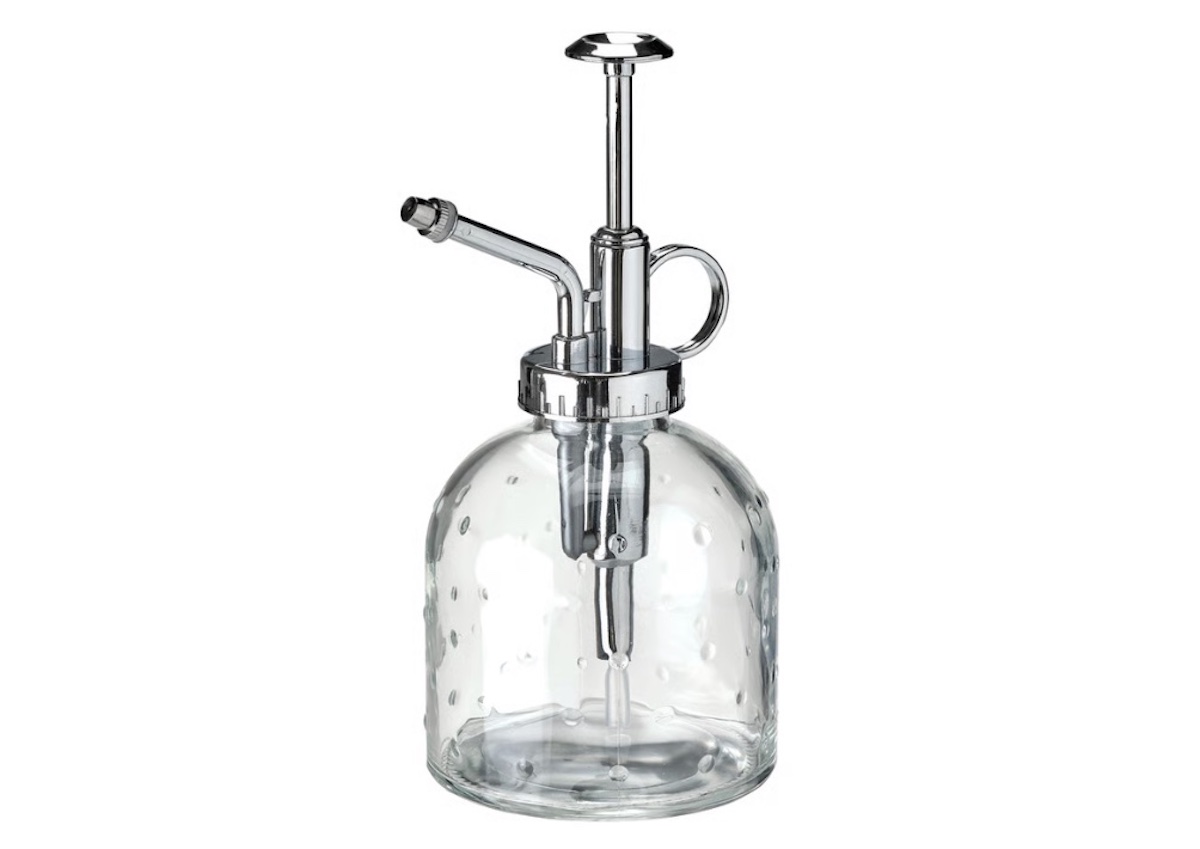 A clear glass plant mister with a metal pump and spout, featuring a round body and a small handle near the neck.