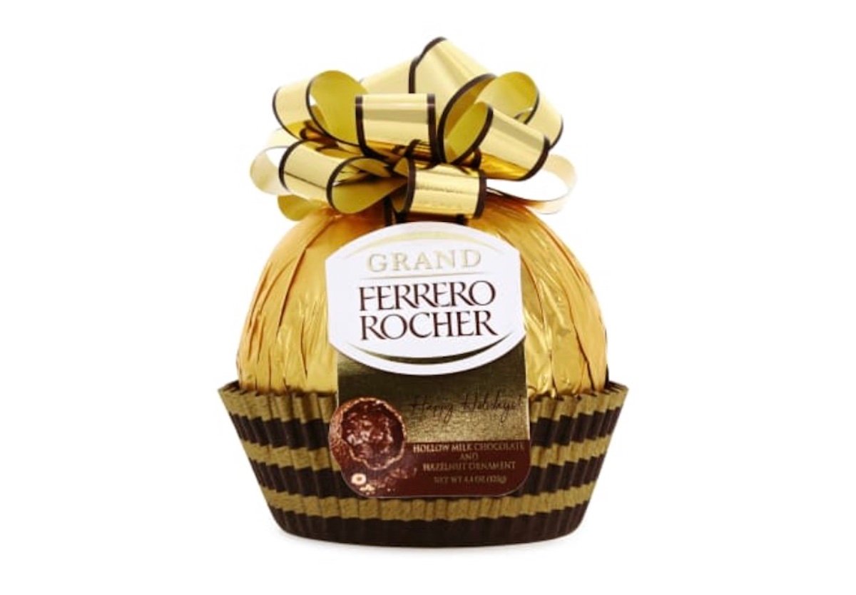A large Ferrero Rocher chocolate wrapped in gold foil, topped with a shiny gold and brown ribbon, and sitting in a brown pleated paper cup. The label reads “Grand Ferrero Rocher.”