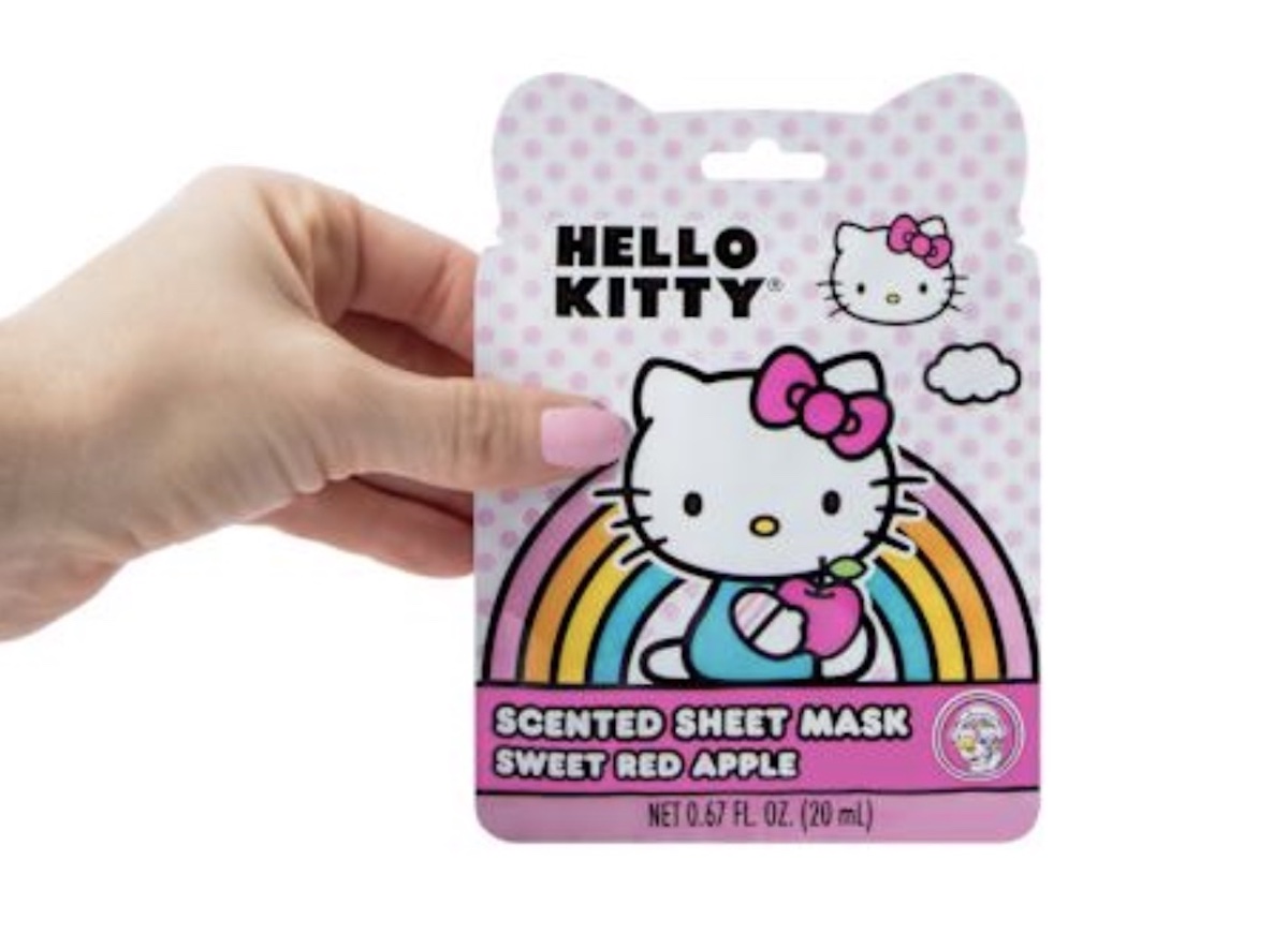 A hand with pink nail polish holds a Hello Kitty scented sheet mask package featuring Hello Kitty, a rainbow, and the text “Scented Sheet Mask Sweet Red Apple.” The packaging is pink with polka dots.