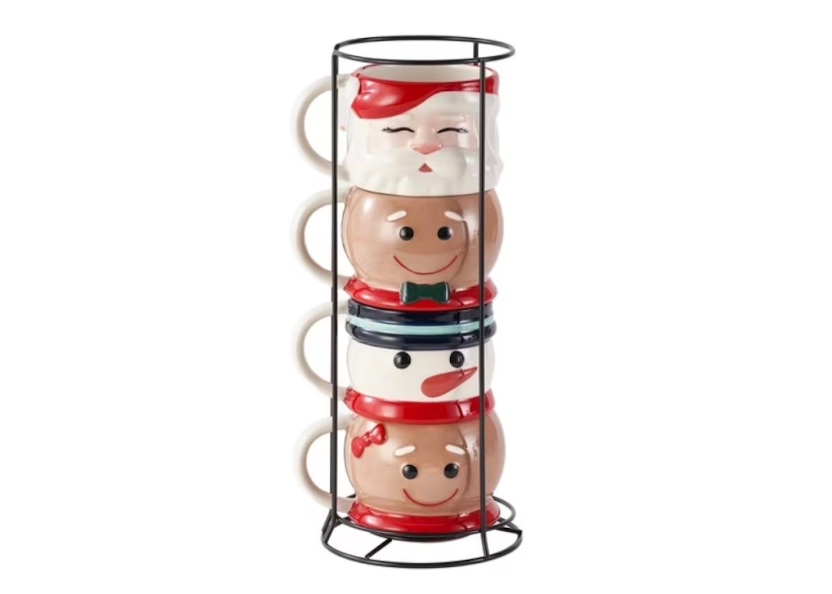 Four festive mugs, each with a different holiday character face—Santa, a gingerbread person, a snowman, and a reindeer—stacked vertically in a black metal holder.