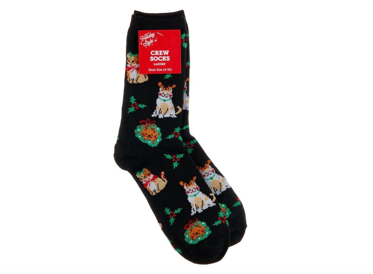 A pair of black crew socks with a festive pattern featuring dogs wearing antlers, holly leaves, and wreaths. A red label on the socks reads "Crew Socks, Ladies, Shoe Size 4-10.