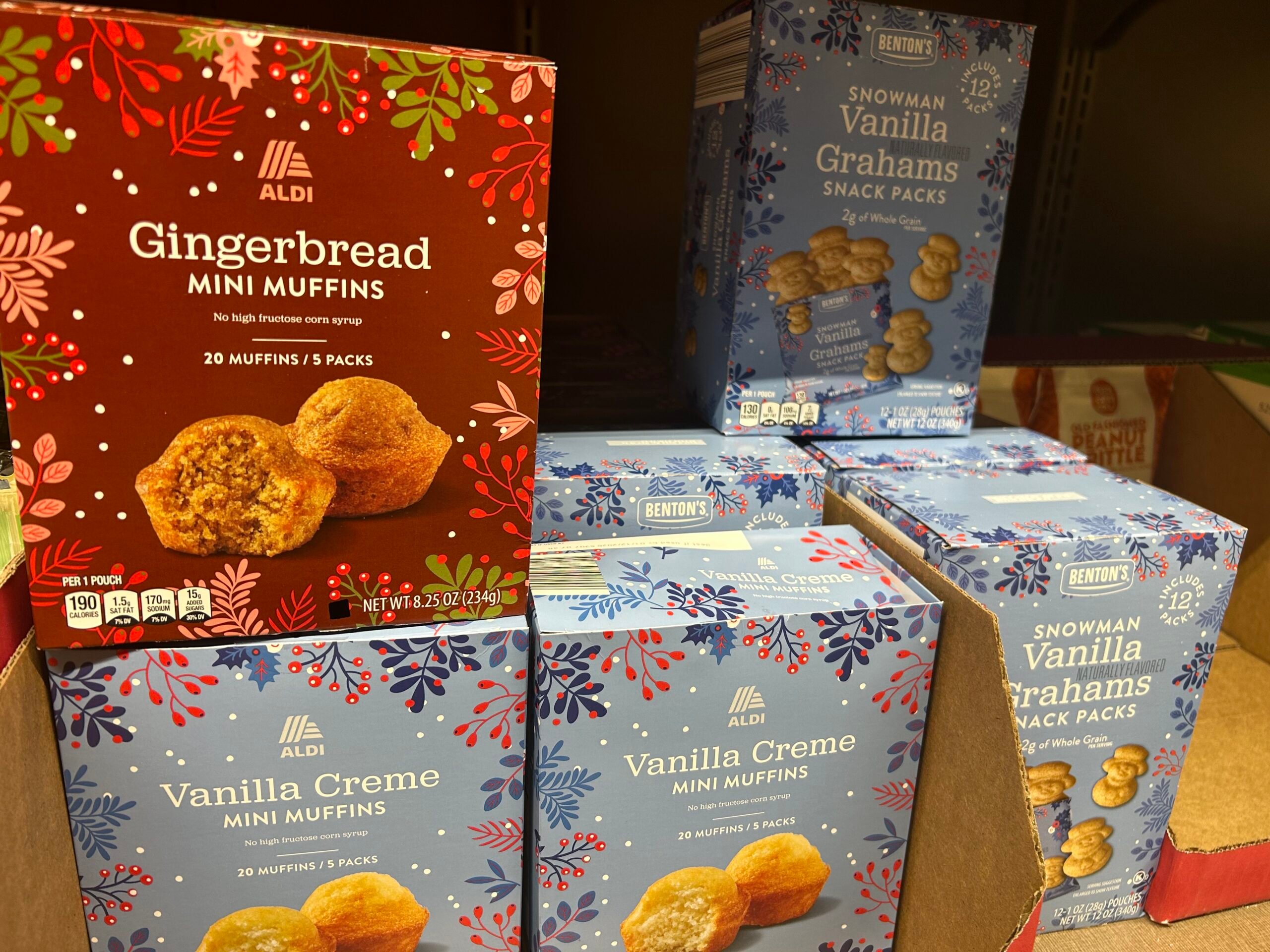 Several boxes of Aldi baked goods on a shelf, including Gingerbread Mini Muffins in a red festive box and Vanilla Creme Mini Muffins and Snowman Vanilla Grahams Snack Packs in blue, snowflake-themed boxes.