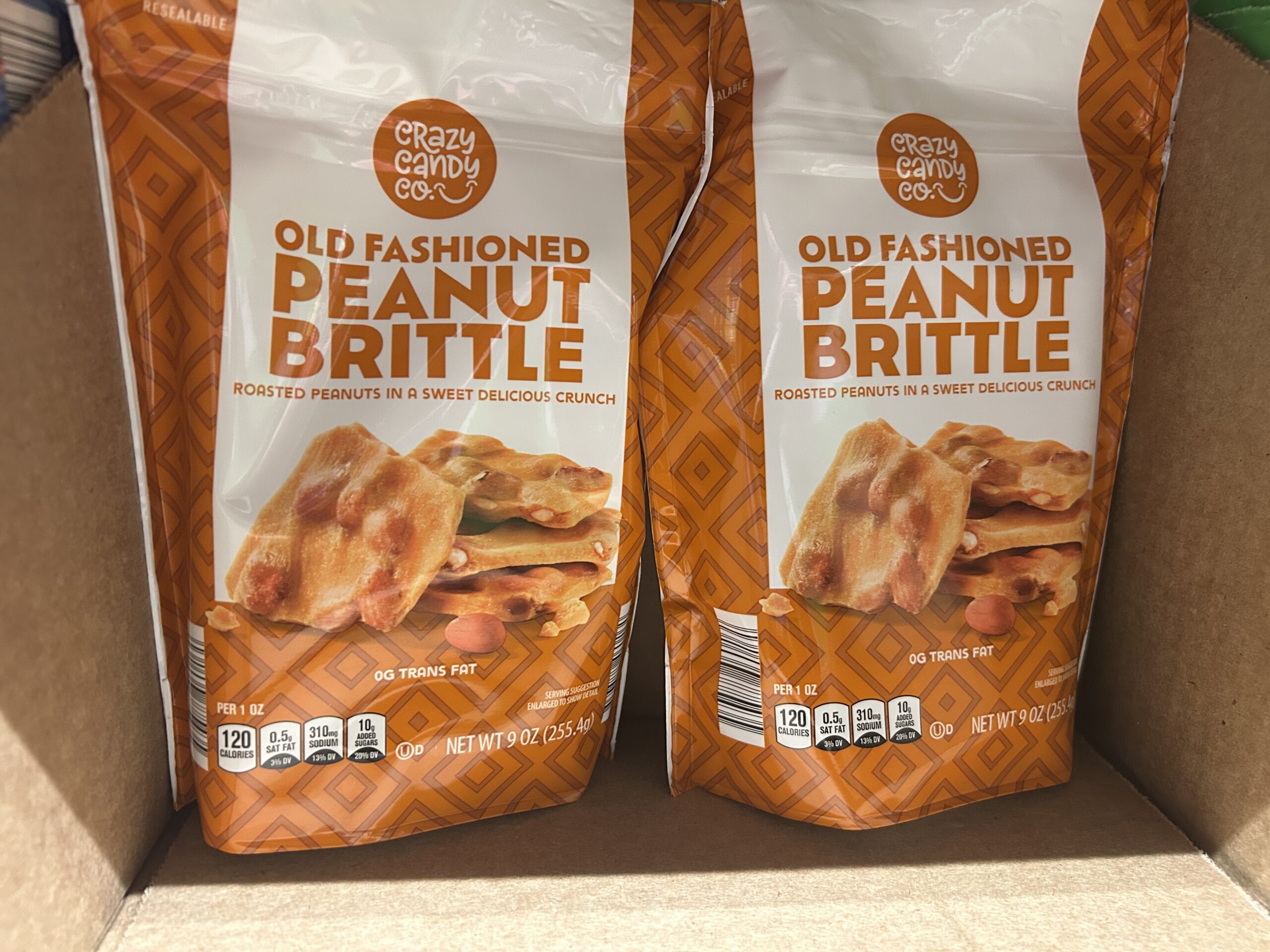 Two bags of Crazy Candy Co. Old Fashioned Peanut Brittle with orange packaging are displayed in a cardboard box. The label highlights roasted peanuts in a sweet, crunchy brittle.