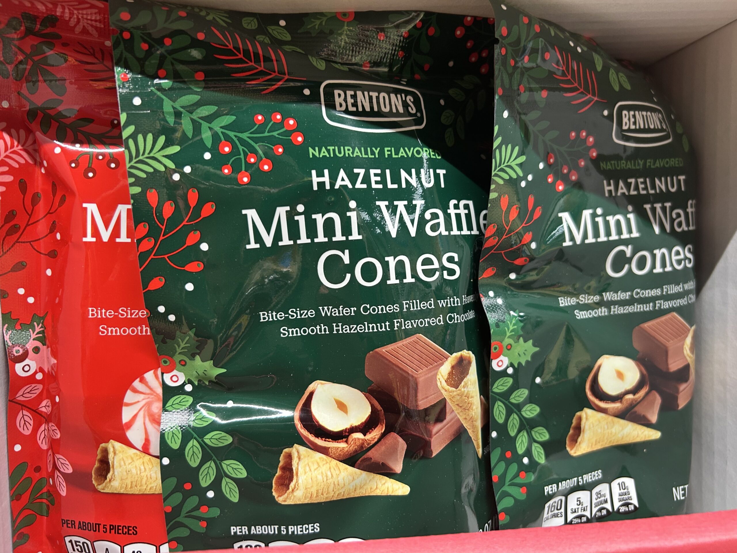 Three bags of Benton's Hazelnut Mini Waffle Cones are displayed on a shelf. The packaging is green with images of waffle cones, chocolate pieces, and hazelnuts, and features festive holiday decorations.