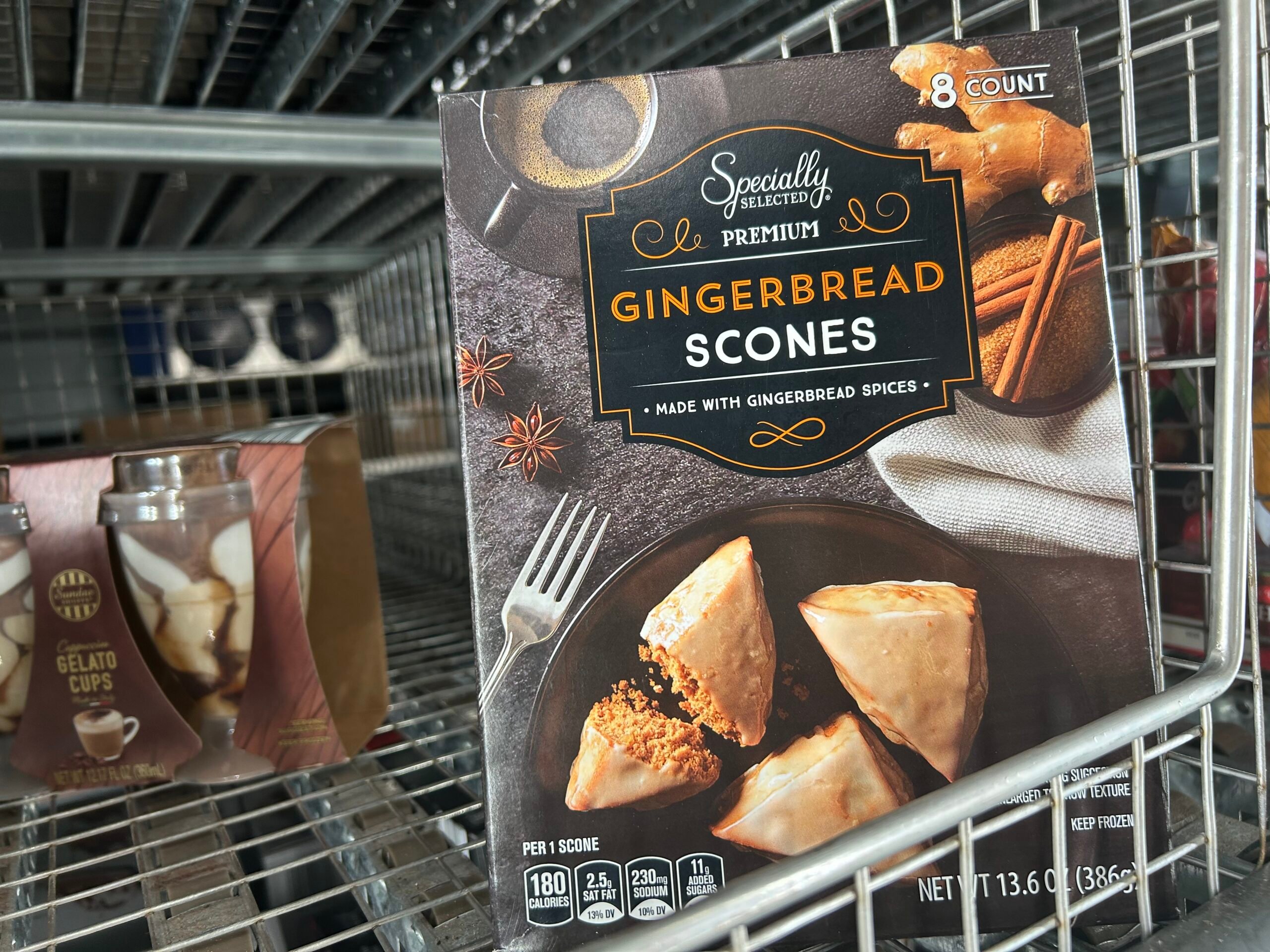 A box of Specially Selected Premium Gingerbread Scones sits on a metal store shelf, displaying an image of the scones on a plate with cinnamon sticks and spices. The box contains 8 scones.