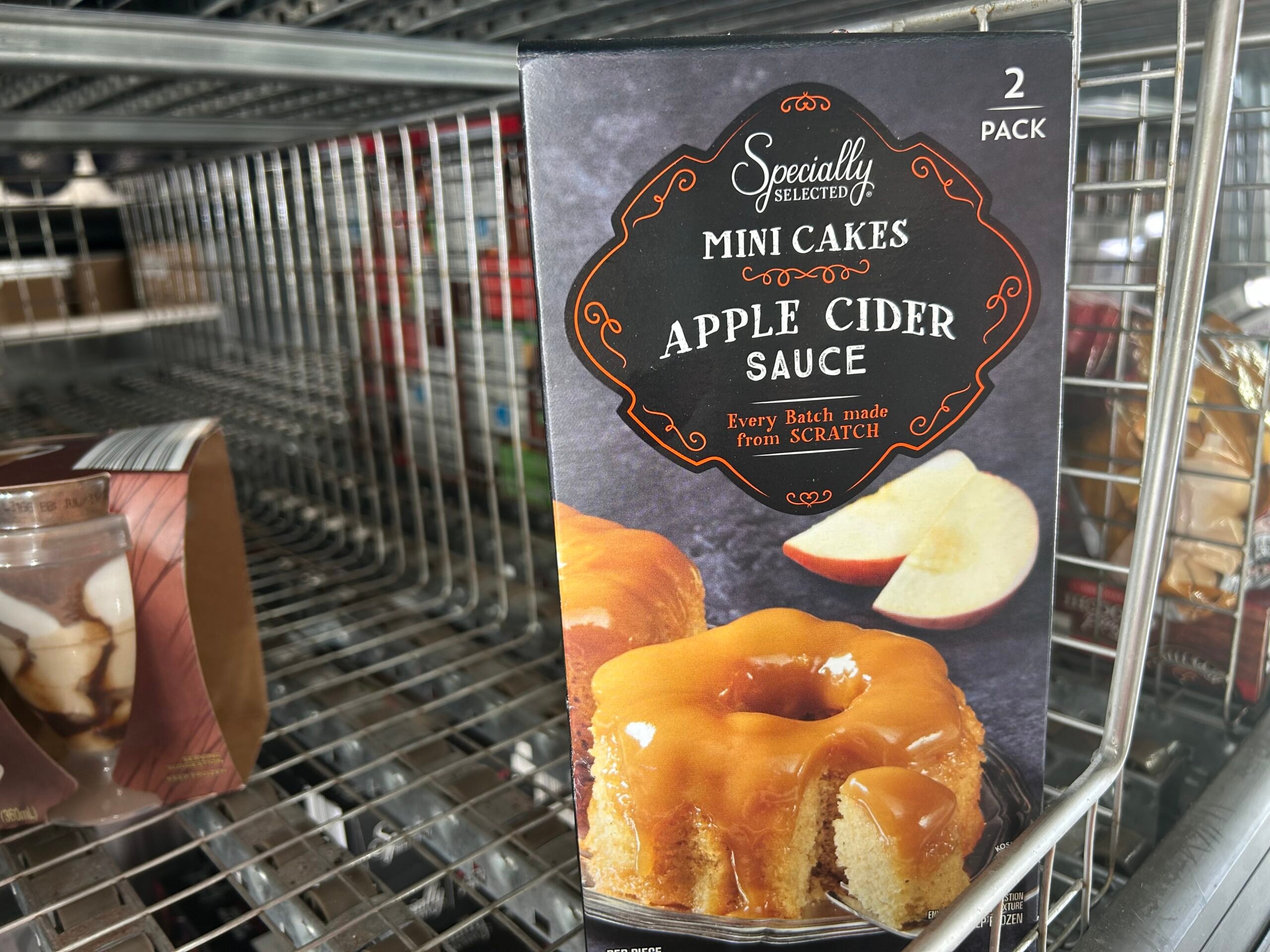 A box labeled "Specially Selected Mini Cakes, Apple Cider Sauce" sits on a metal shelf. The box shows mini cakes topped with sauce and apple slices. The packaging says “2 pack” and “Every batch made from scratch.”