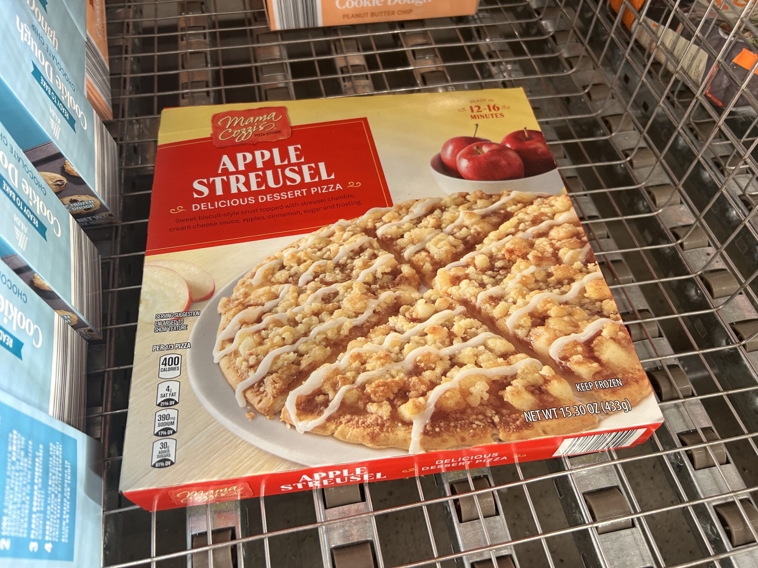 A box of Mama Cozy’s Apple Streusel Delicious Dessert Pizza sits on a store shelf. The packaging features an image of the dessert pizza topped with crumble and apples. The box weighs 15.33 oz (435g).
