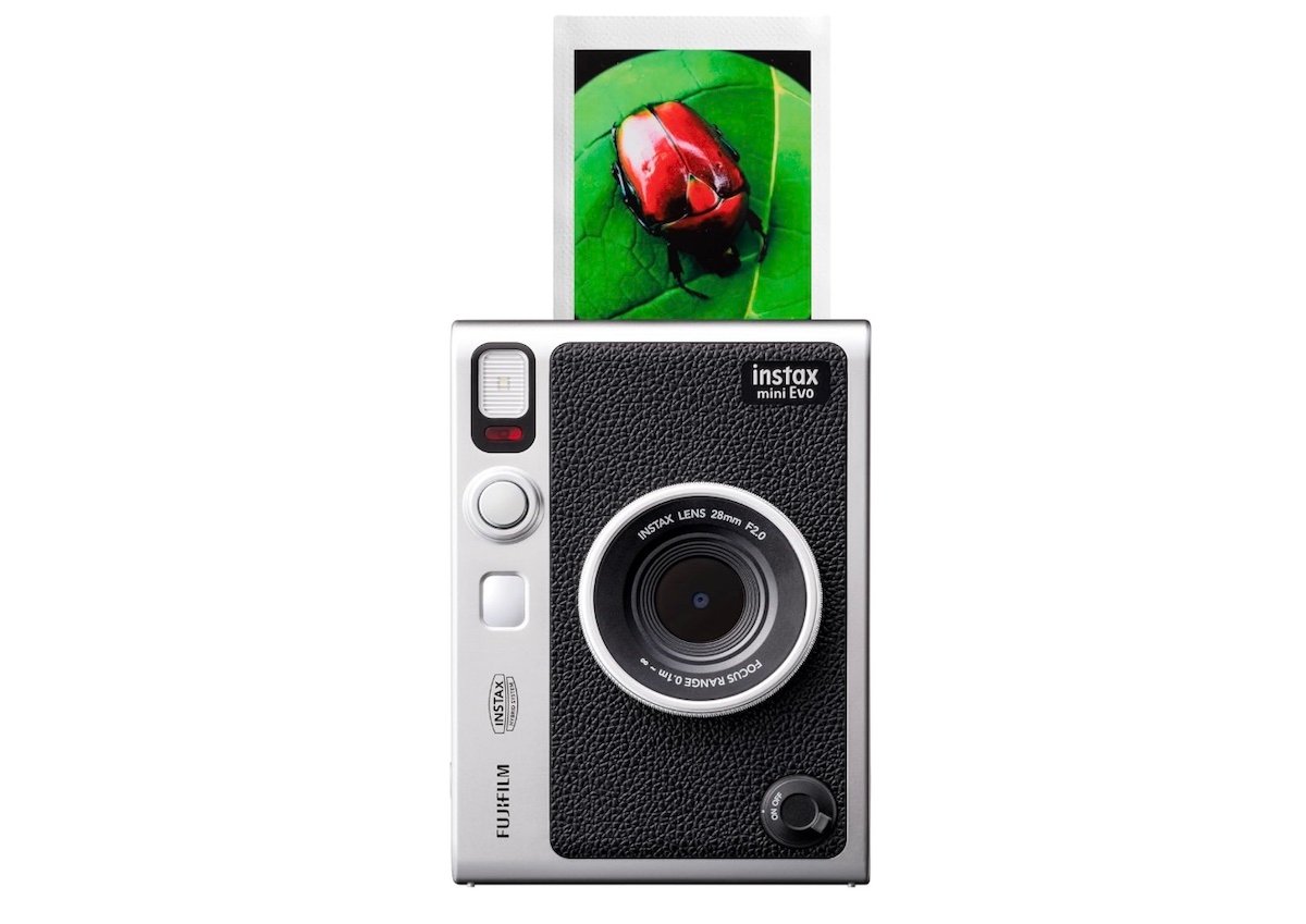 A Fujifilm Instax Mini Evo instant camera prints a photo of a red beetle on a green leaf. The camera is black and silver with a textured surface and a developed photo emerging from the top.