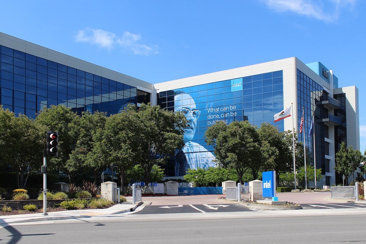 Modern glass office building with a large mural and the words "What can be done, can be outdone." Trees and flags stand in front, along with a blue NTT sign near the street under a blue sky.