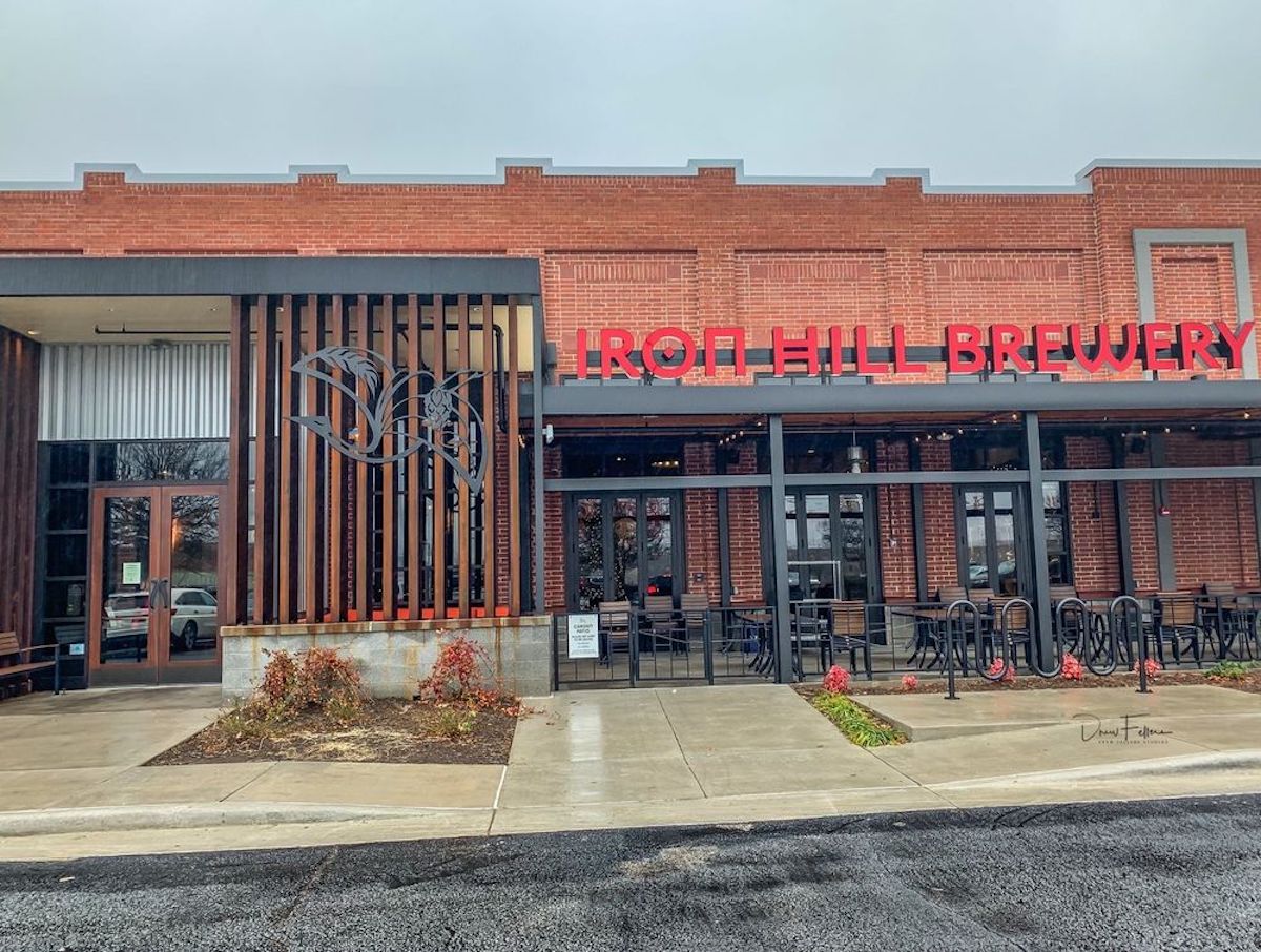 A brick building with large red letters reading "IRON HILL BREWERY" above black-framed windows. There is a metal decorative design and outdoor seating in front of the entrance on a cloudy day.