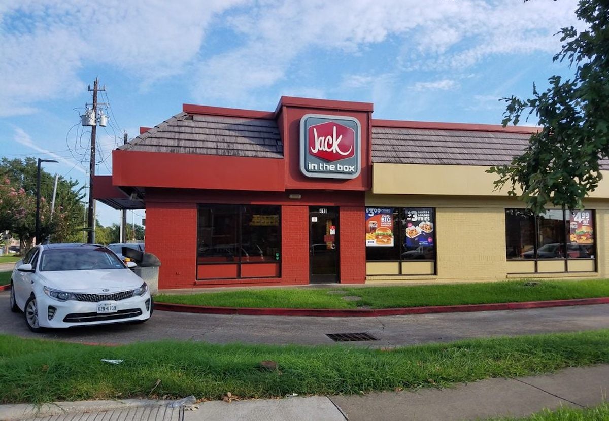 A white car is parked in front of a Jack in the Box fast food restaurant with a red, brown, and beige exterior. The sky is partly cloudy, and there is green grass and a sidewalk in the foreground.