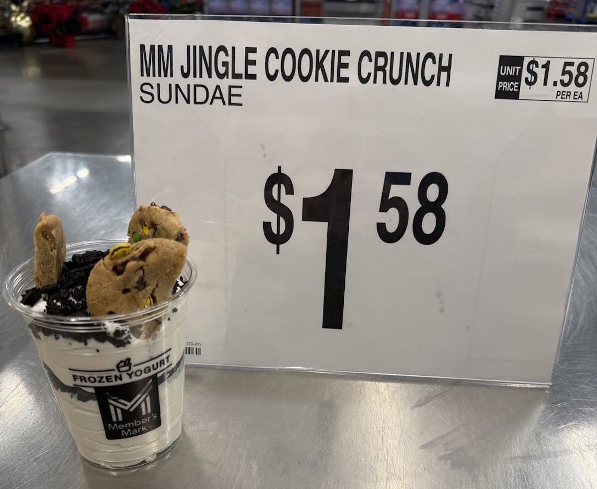 A cup of frozen yogurt topped with cookies and candy pieces sits on a counter next to a sign that reads "MM Jingle Cookie Crunch Sundae $1.58.
