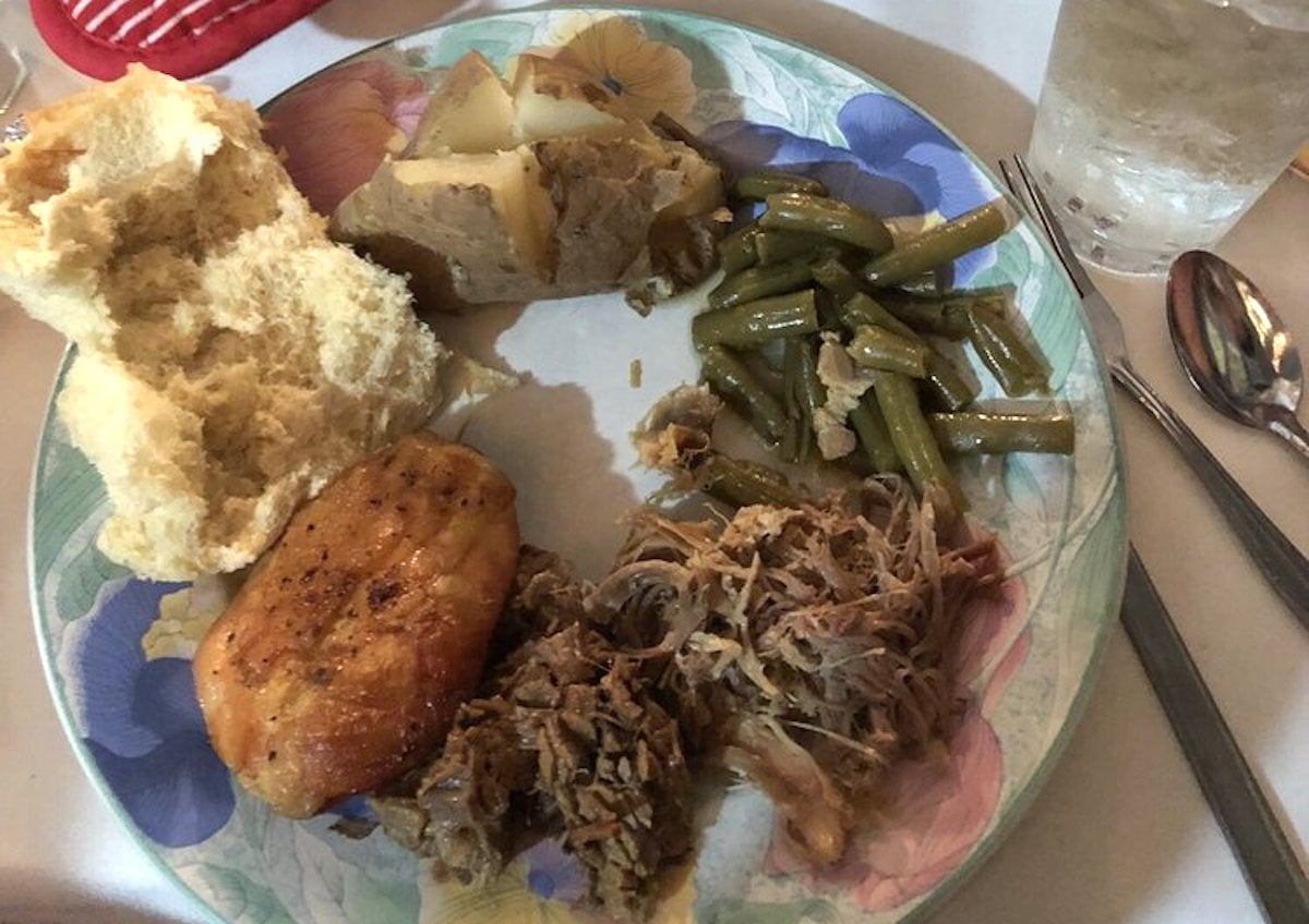 A plate from one of the city's under-the-radar restaurants features a biscuit, baked chicken, shredded meat, green beans, a slice of potato with meat, and a glass of iced water. Silverware rests neatly beside the plate.