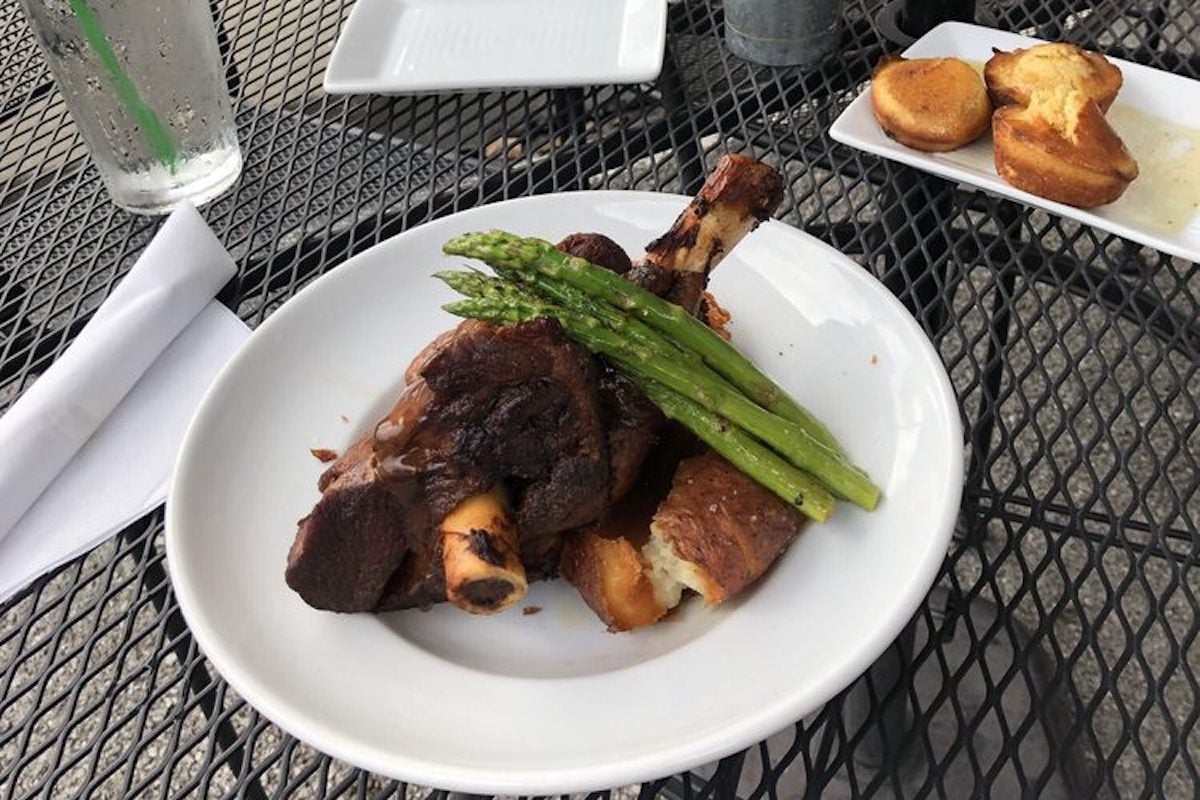 A white plate with braised short ribs on bones, garnished with grilled asparagus and bread—just the kind of cozy meal you’d find at under-the-radar restaurants. Nearby sit a napkin, a glass of water with a straw, and cornbread muffins with butter.